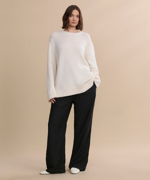 A woman stands against a beige background wearing the Cashmere Amelia Crewneck, paired with wide-leg black pants and white shoes, with one hand in her pocket and a relaxed expression.