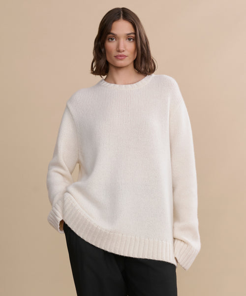 A woman with wavy brown hair wears the oversized ivory Cashmere Amelia Crewneck sweater and loose black pants, standing against a plain beige background.