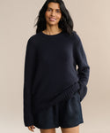 A woman with long dark hair wears the Cashmere Amelia Crewneck and matching shorts, standing against a plain beige background and smiling softly at the camera.