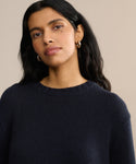 A woman with long dark hair and gold hoop earrings wears the Cashmere Amelia Crewneck in navy and looks slightly to the side against a neutral beige background.