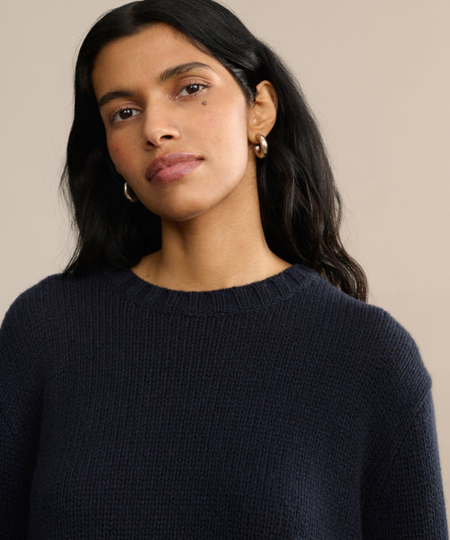 A woman with long dark hair and gold hoop earrings wears the Cashmere Amelia Crewneck in navy and looks slightly to the side against a neutral beige background.