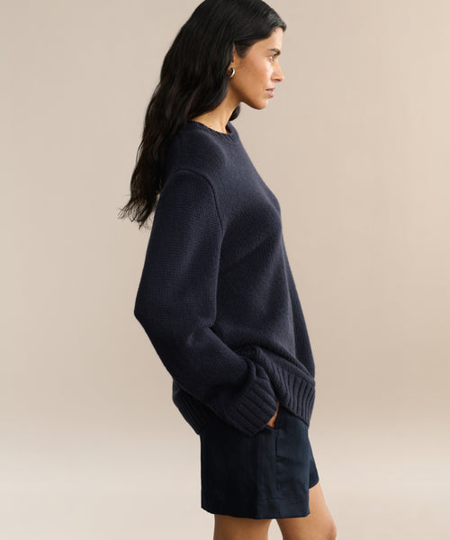A woman with long dark hair stands in profile against a neutral background, wearing the oversized navy Cashmere Amelia Crewneck sweater with long sleeves and dark shorts.