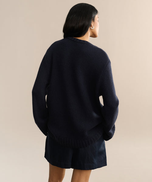 A person with long dark hair, wearing the Cashmere Amelia Crewneck in navy and dark shorts, stands facing away from the camera against a neutral beige background.