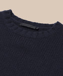 Close-up of the Cashmere Amelia Crewneck, a navy blue chunky knit sweater with a round neckline, displayed on a beige background. The black clothing label is visible inside the collar.