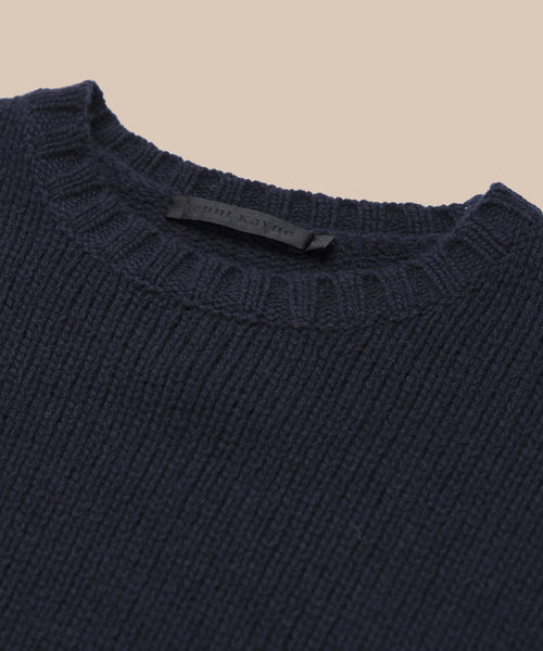 Close-up of the Cashmere Amelia Crewneck, a navy blue chunky knit sweater with a round neckline, displayed on a beige background. The black clothing label is visible inside the collar.