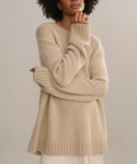 A person with curly hair wears the Cashmere Amelia Crewneck, a loose, lightweight beige sweater with long sleeves, standing against a neutral background and resting one hand on their chin.