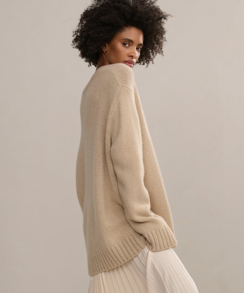 A woman with curly hair looks over her shoulder while wearing the Cashmere Amelia Crewneck and a light pleated skirt, standing against a neutral background.