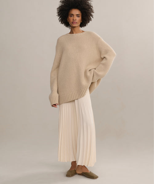 A woman with curly hair wears the Cashmere Amelia Crewneck sweater, paired with a long pleated cream skirt and brown flats, standing against a plain light backdrop.