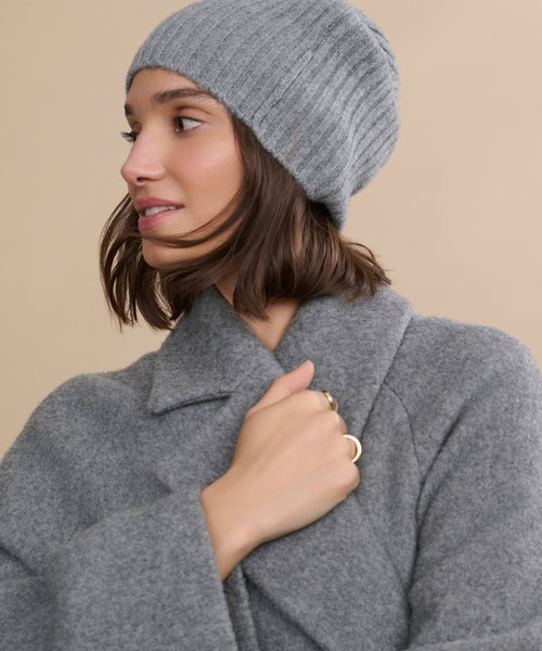 A woman with shoulder-length brown hair wears a cozy grey Cashmere Beanie and matching coat, looking to the side. She holds her lapel, displaying gold rings. The beige background emphasizes the beanie’s stylish comfort.