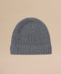 The Cashmere Beanie is a grey ribbed knit hat with a folded brim, made from lightweight cashmere and shown laid flat on a light beige background.