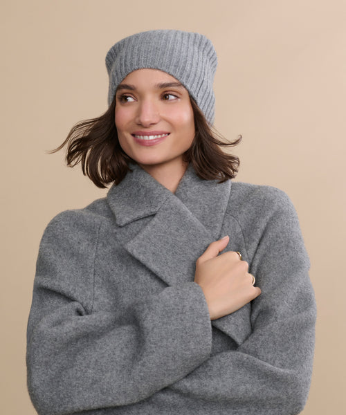 A woman in a gray wool coat and gray knit hat smiles with her arms crossed before a beige background, the cozy Cashmere Beanie adding the perfect touch of style to her chic, layered look.