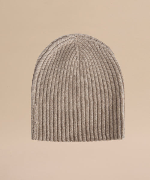 The Cashmere Beanie, featuring a beige ribbed knit design, is displayed upright against a plain light tan background.