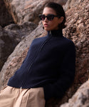 A person wearing dark sunglasses, the Boyd Cashmere Sweater in navy, and beige pants leans against large rocks outdoors, looking off to the side with a neutral expression.