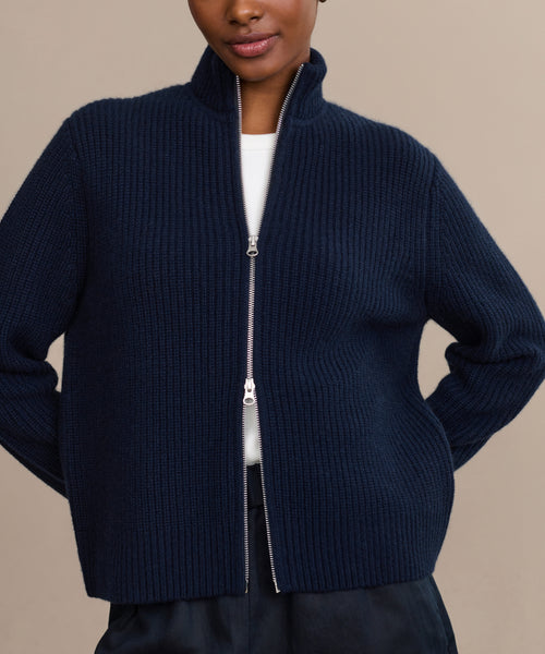A person wearing the Boyd Cashmere Sweater, a dark navy blue fisherman rib zip-up with a high collar, layered over a white top and dark pants, stands hands on hips against a beige background.