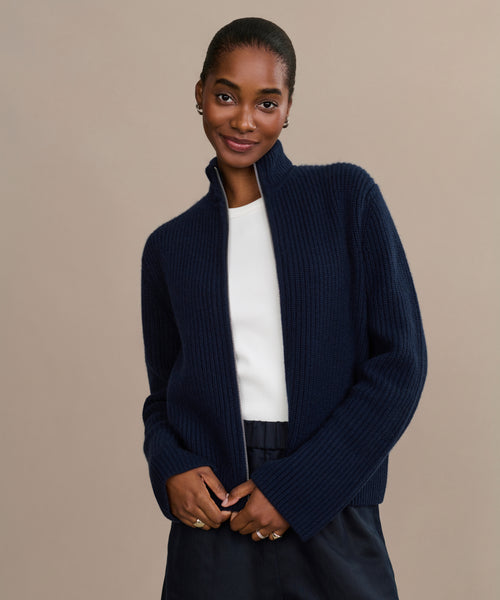 A woman stands against a beige background, smiling softly in the Boyd Cashmere Sweater—a navy blue mock neck with fisherman rib texture—layered over a white top and dark pants, gently holding the sweater near her waist.