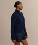 A woman stands sideways against a beige background, wearing a navy Boyd Cashmere Sweater with a zip-up mock neck and dark shorts. Her hair is pulled back, and her arms rest by her sides.