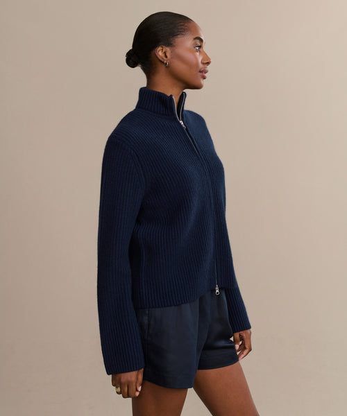 A woman stands sideways against a beige background, wearing a navy Boyd Cashmere Sweater with a zip-up mock neck and dark shorts. Her hair is pulled back, and her arms rest by her sides.