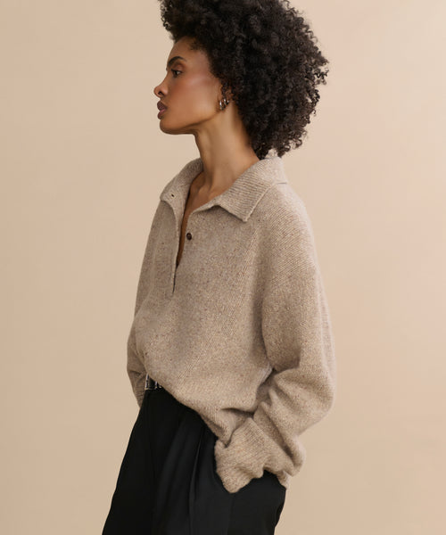 A woman with short curly hair poses in profile against a beige background, wearing the oversized Cashmere Burke Polo sweater with buttons and black pants, her hands casually tucked in her pockets.