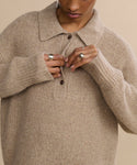 A person is buttoning the top button of the Cashmere Burke Polo in beige, shown from shoulders to just above the lips, and wearing several silver rings.