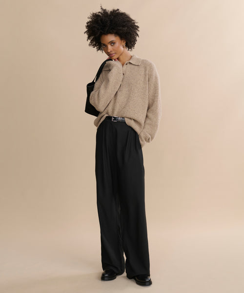 A person with short curly hair wears the oversized Cashmere Burke Polo in beige, wide-leg black pants, and black shoes, carrying a black shoulder bag against a plain beige background.