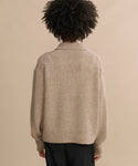 Person with curly hair wears the Cashmere Burke Polo in beige and black pants, standing with their back to the camera against a plain tan background.