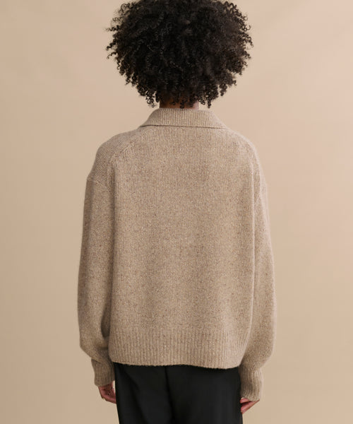 Person with curly hair wears the Cashmere Burke Polo in beige and black pants, standing with their back to the camera against a plain tan background.