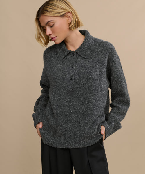 A woman with short blonde hair wears the Cashmere Burke Polo and black pants, standing against a plain beige background with her hands in her pockets, looking down.