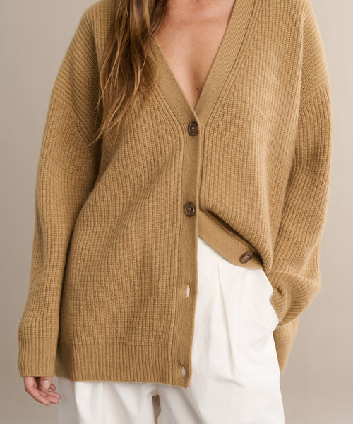 A person wears the Cashmere Cocoon Cardigan—a slouchy, tan ribbed knit with brown buttons—partially unbuttoned over high-waisted, wide-leg white pants, with one hand tucked in their pocket.