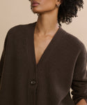 A woman with short curly hair wears the oversized Cashmere Cocoon Cardigan in dark brown, featuring a deep V-neck and single button. She stands against a neutral beige background, her face partially out of frame.