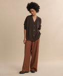 A person stands against a beige background wearing the oversized Cashmere Cocoon Cardigan in dark brown, rust wide-leg trousers, and brown shoes, with one hand in their pocket and looking slightly downward.
