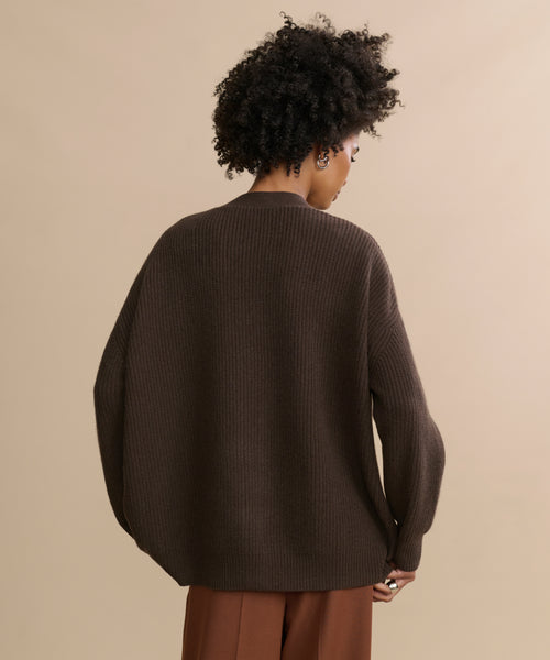 A person with short curly hair is seen from behind wearing the Cashmere Cocoon Cardigan in dark brown and matching brown pants, standing against a plain beige background.