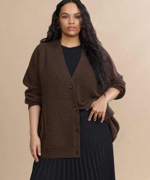 A woman with long wavy hair models the oversized dark brown Cashmere Cocoon Cardigan over a black top and pleated black skirt, standing against a plain beige background.