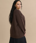 Woman with long, wavy dark hair wearing the Cashmere Cocoon Cardigan in brown and a black pleated skirt stands sideways against a beige background, looking toward the camera.