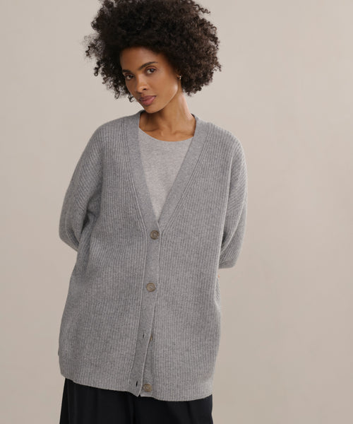 A person with curly hair wears the Cashmere Cocoon Cardigan, a slouchy gray button-up, layered over a light gray shirt, standing hands behind their back against a plain light background.