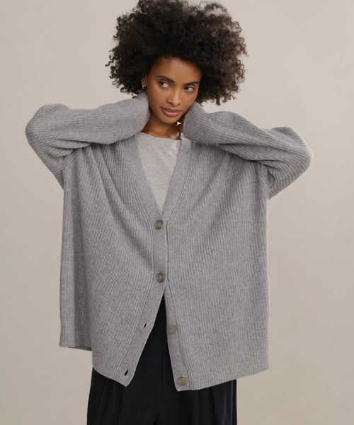 A person with curly hair wears the loose, gray Cashmere Cocoon Cardigan over a light shirt and black pants, standing with both hands touching the back of their neck against a plain background.