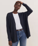 A person wears the Cashmere Cocoon Cardigan in dark blue over a white t-shirt and blue jeans, standing with one hand on their head against a plain background.