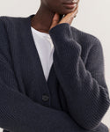 A person wearing the Cashmere Cocoon Cardigan in dark blue over a white shirt rests their chin on their hand, partially covering their face. The cardigan features a ribbed texture and visible button detail.