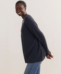 A smiling woman stands sideways with her arms behind her back against a plain, light background, wearing blue jeans and the dark gray Cashmere Cocoon Cardigan.