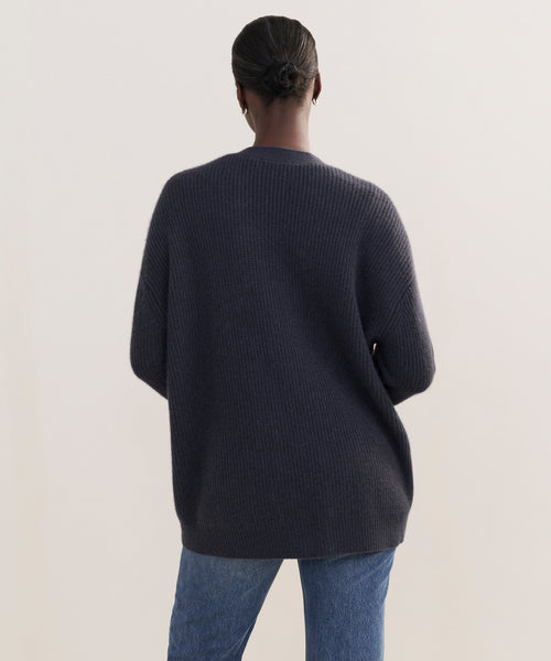 A person with dark hair in a low bun is seen from behind, wearing the Cashmere Cocoon Cardigan and blue jeans, standing against a plain light background.