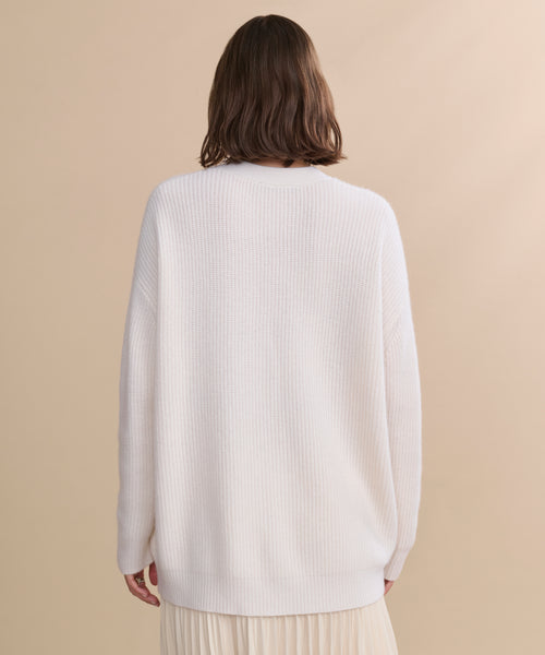 A person with shoulder-length brown hair stands with their back to the camera, wearing the Cashmere Cocoon Cardigan in textured white knit against a neutral beige background.