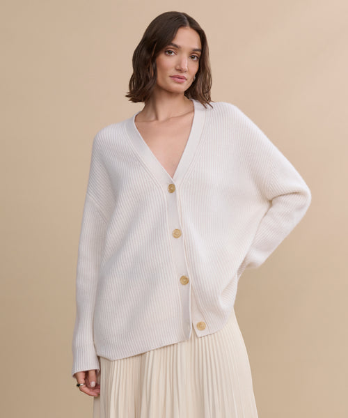 A woman with shoulder-length brown hair models the Cashmere Cocoon Cardigan, featuring large buttons, layered over a matching pleated skirt against a plain beige background.