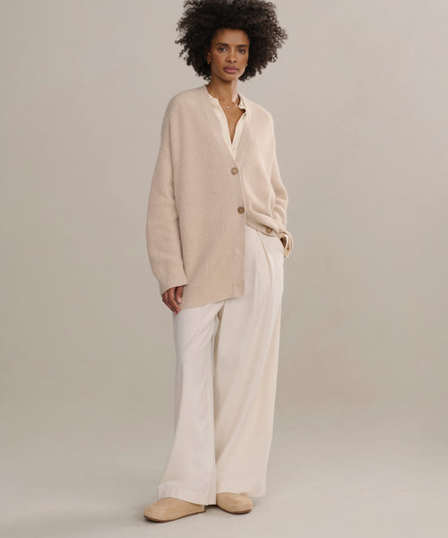 Wearing the Cashmere Cocoon Cardigan in beige over a white shirt, wide-leg white pants, and beige slippers, a person with short curly hair stands against a plain light background.