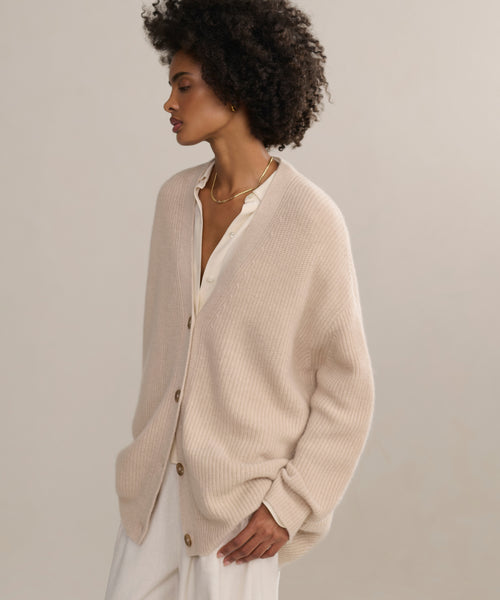 A woman with short curly hair models the Cashmere Cocoon Cardigan over a white blouse and pants, hands in pockets, gazing to the side against a neutral background.
