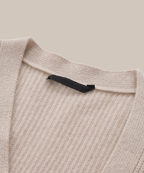 Close-up of the Cashmere Cocoon Cardigan, featuring beige ribbed knit fabric, a V-neckline, and a black label at the back of the neck. The background is solid light beige.
