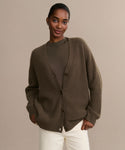 A woman stands against a neutral background wearing the oversized Cashmere Cocoon Cardigan over a matching brown top and white pants, hands in pockets and with a relaxed expression.