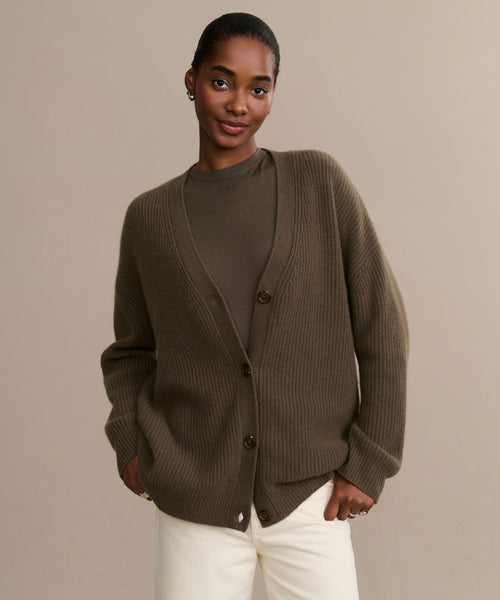 A woman stands against a neutral background wearing the oversized Cashmere Cocoon Cardigan over a matching brown top and white pants, hands in pockets and with a relaxed expression.