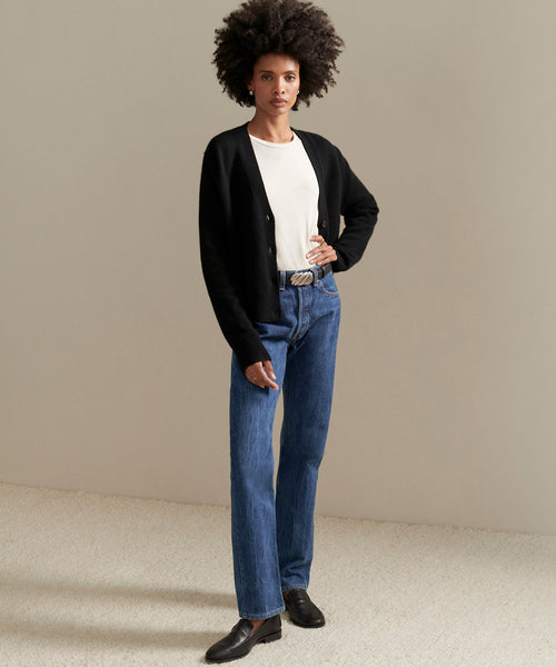 A person with short curly hair stands indoors against a plain background, wearing the black Cashmere Colette Cardigan over a white t-shirt, blue relaxed-fit jeans, a black belt, and black shoes, with one hand in their pocket.