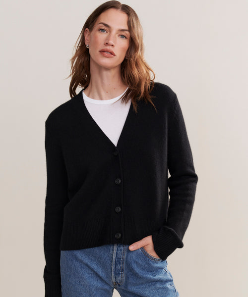 A woman with light brown hair wears the Cashmere Colette Cardigan in black, styled over a white t-shirt and blue jeans, standing against a plain light background with one hand in her pocket.