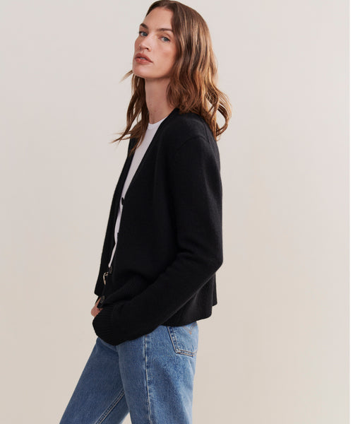 A person with long brown hair models the relaxed fit Cashmere Colette Cardigan in black, layered over a white top and blue jeans, standing sideways and facing the camera against a plain light background.