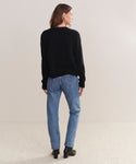 A person with light brown hair stands facing away from the camera against a neutral background, wearing the black Cashmere Colette Cardigan in a relaxed fit, paired with blue jeans and black ankle boots.